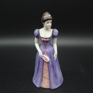 Może przedstawiać: Porcelanowa figurka kobiety w fioletowej i różowej sukni z białym koronkowymi kołnierzykiem i koroną. Trzyma wachlarz w prawej ręce.