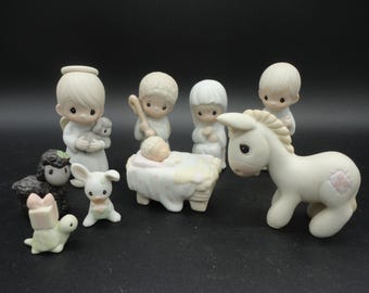 Może przedstawiać: Zestaw ceramicznych figurek przedstawiających scenę Bożego Narodzenia. Zestaw zawiera figurki anioła, Dzieciątka Jezus w żłóbku, osła i innych zwierząt. Figurki są głównie białe z pastelowymi akcentami.