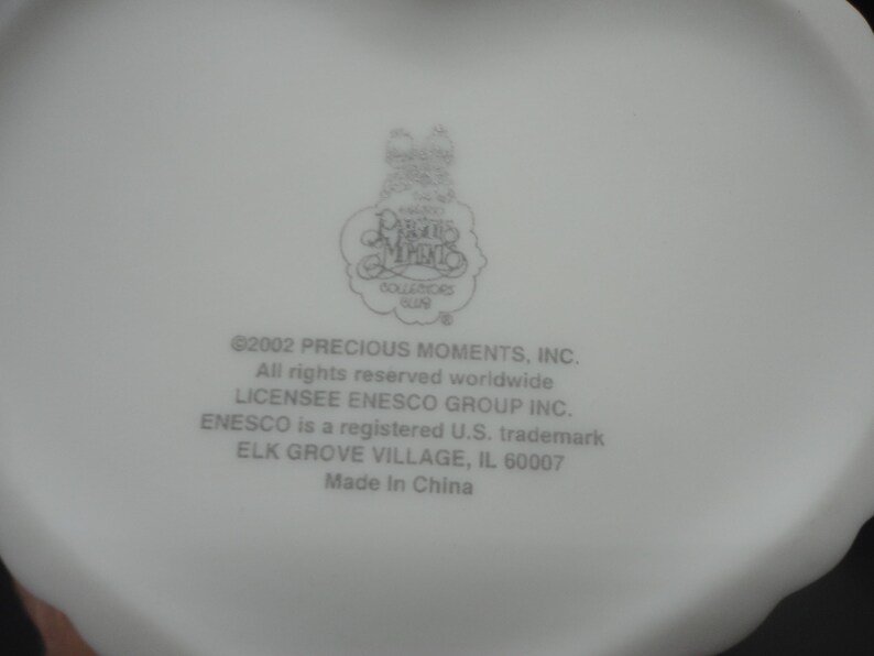 Puede incluir: Una base de cer&aacute;mica blanca con el logotipo de Precious Moments Collectors Club y el texto: 2002 PRECIOUS MOMENTS, INC. All rights reserved worldwide. LICENSEE ENESCO GROUP INC. ENESCO is a registered U.S. trademark. ELK GROVE VILLAGE, IL 60007. Made In China.