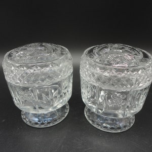 Op de afbeelding: Twee glazen potten met deksel, gemaakt van helder glas met een ruitpatroon en bloemenmotief. De potten hebben een voet.