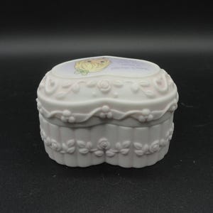 Puede incluir: Un joyero de porcelana blanca en forma de coraz&oacute;n con acabado mate y detalles en relieve de rosas y hojas en los laterales. La tapa muestra una ilustraci&oacute;n colorida de un personaje rubio con un lazo verde y texto impreso.
