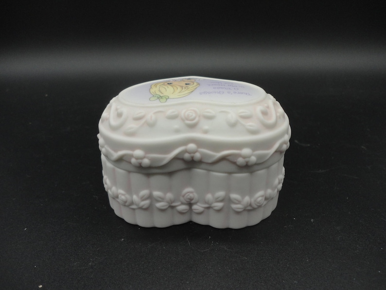 Puede incluir: Un joyero de porcelana blanca en forma de coraz&oacute;n con acabado mate y detalles en relieve de rosas y hojas en los laterales. La tapa muestra una ilustraci&oacute;n colorida de un personaje rubio con un lazo verde y texto impreso.