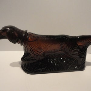 Può includere: Una bottiglia di vetro marrone a forma di cane, probabilmente un segugio, con una superficie testurizzata. La bottiglia è vuota e ha un tappo.