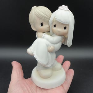 Puede incluir: Figura de boda de cerámica que representa a un novio llevando a una novia. El novio tiene el pelo claro, y la novia lleva un vestido blanco y un velo. La figura está sobre una base blanca y tiene un acabado mate. Altura aproximada de 15 cm.
