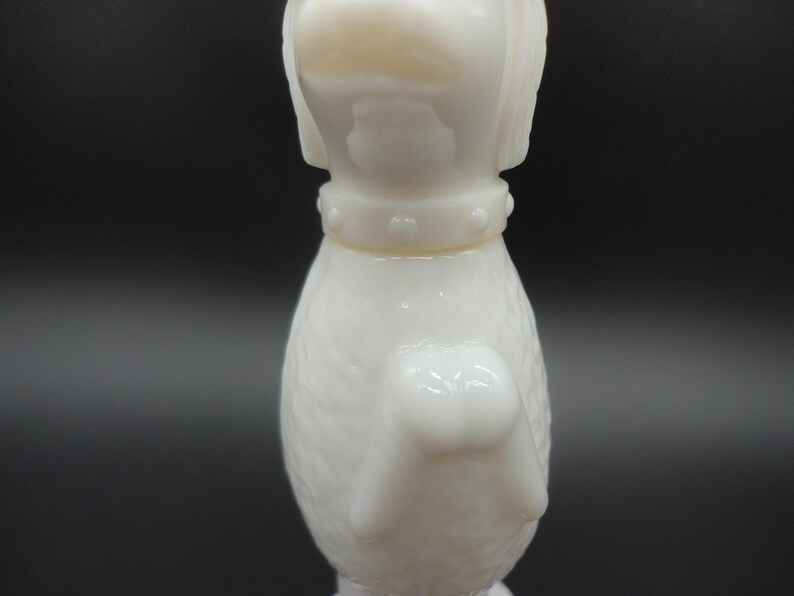 May include: A white, vintage dog figurine with a textured body and a collar with small, raised dots. The dog's head is upright, and the figurine has a glossy finish. The background is a dark gray.