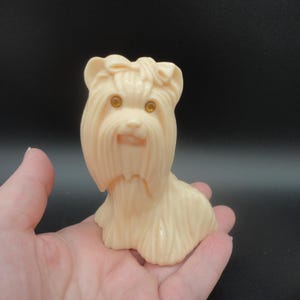 May include: A light beige plastic figurine of a Yorkshire Terrier dog. The dog has detailed fur, a bow on its head, and amber-colored eyes. The figurine is held in a hand against a dark background.