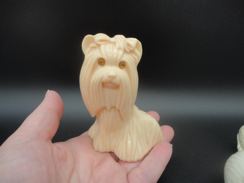 May include: A light beige plastic figurine of a Yorkshire Terrier dog. The dog has detailed fur, a bow on its head, and amber-colored eyes. The figurine is held in a hand against a dark background.