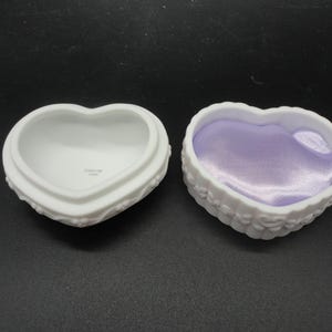 Puede incluir: Dos cajas de baratijas de porcelana blanca en forma de coraz&oacute;n con detalles florales en relieve en los lados exteriores. Una caja est&aacute; vac&iacute;a con un interior blanco liso, mientras que la otra est&aacute; forrada con un inserto de tela de sat&eacute;n lavanda brillante.