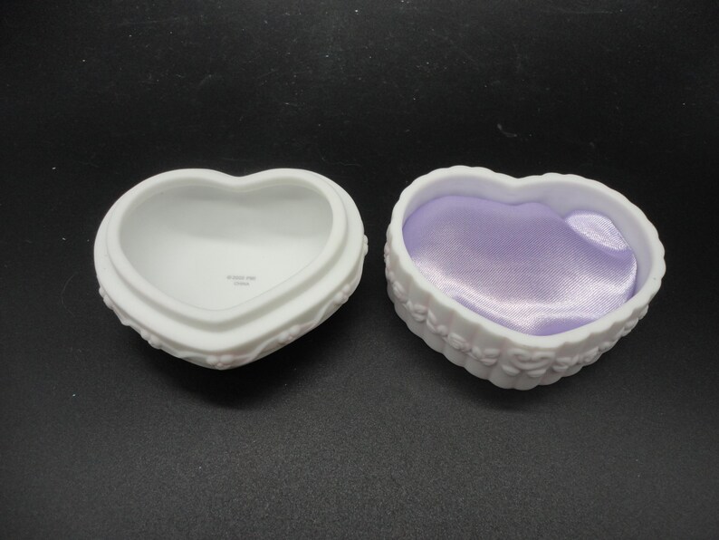 Puede incluir: Dos cajas de baratijas de porcelana blanca en forma de coraz&oacute;n con detalles florales en relieve en los lados exteriores. Una caja est&aacute; vac&iacute;a con un interior blanco liso, mientras que la otra est&aacute; forrada con un inserto de tela de sat&eacute;n lavanda brillante.