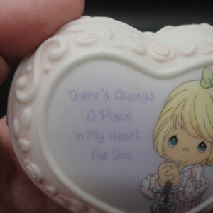 Puede incluir: Un joyero de porcelana en forma de coraz&oacute;n con un borde floral en relieve. La parte frontal presenta una ilustraci&oacute;n de dibujos animados de un ni&ntilde;o rubio con un lazo verde y el texto morado: There's Always A Place In My Heart For You.
