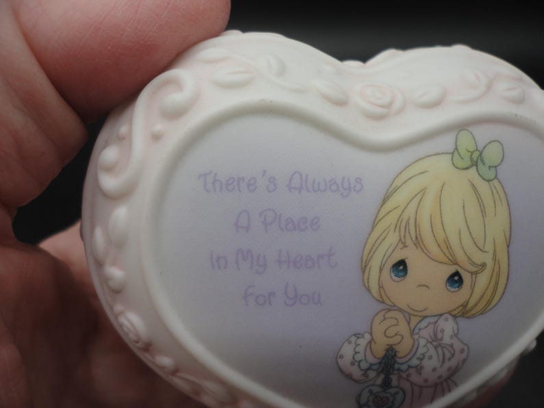 Puede incluir: Un joyero de porcelana en forma de coraz&oacute;n con un borde floral en relieve. La parte frontal presenta una ilustraci&oacute;n de dibujos animados de un ni&ntilde;o rubio con un lazo verde y el texto morado: There's Always A Place In My Heart For You.
