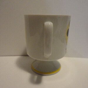 May include: White ceramic mug with a yellow and black smiley face design. The mug has a handle and a pedestal base.