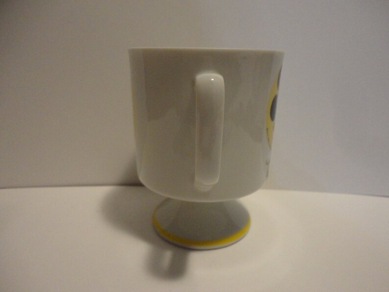 May include: White ceramic mug with a yellow and black smiley face design. The mug has a handle and a pedestal base.