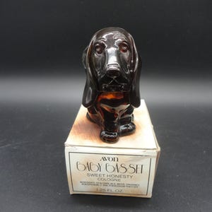 May include: A brown glass cologne bottle in the shape of a Basset Hound dog, presented on a box. The box is white and features the text "AVON BABY BASSET SWEET HONESTY COLOGNE 1.25 FL OZ."