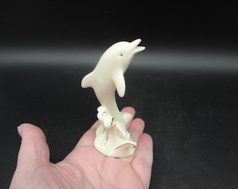 May include: A white porcelain figurine of a dolphin leaping out of the water. The dolphin is painted with gold accents and is standing on a base that resembles a wave.