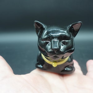 May include: A small, glossy black cat figurine with pointed ears and closed eyes. The cat has a smooth, reflective surface and wears a simple, solid yellow collar around its neck. The figurine is sculpted in a seated position with a rounded head.
