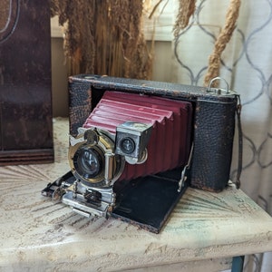 Könnte beinhalten: Eine vintage, schwarze, lederbezogene Faltenkamera mit einem roten Balg und einem silbernen Objektiv. Die Kamera ist geöffnet und der Balg ist ausgefahren.