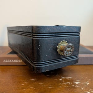 May include: A rectangular dark brown wooden box with horizontal grooved detailing along the sides. The front features a circular brass knob with an ornate, scalloped backplate. The box is positioned on a flat, brown rectangular base.