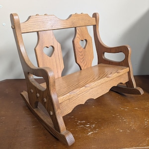 May include: A small wooden rocking bench featuring a natural light brown wood grain finish. The backrest includes two vertical slats, each with a heart-shaped cutout. The bench has curved armrests and a scalloped apron below the seat.