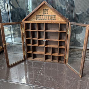May include: A wooden display cabinet with a gabled top and glass doors. The cabinet has multiple small square compartments inside. The words "MY COLLECTION" are printed in gold lettering on the top. The cabinet is open, revealing the interior.