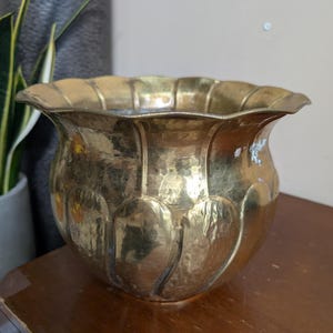 Puede incluir: Una maceta de latón pulido con un borde festoneado en forma de pétalos y un cuerpo redondeado y bulboso con hendiduras verticales. La superficie metálica muestra un tono dorado cálido y reflectante con variaciones de textura.