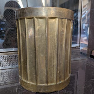 May include: A cylindrical brass container with vertical fluted sides. The container has a rounded base and a thick, flat rim. The brass has a warm, aged patina. The container is likely a decorative item or a storage vessel.