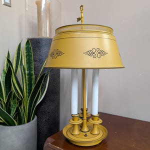 May include: A yellow metal table lamp featuring a wide, conical shade with black decorative leaf patterns. The lamp base holds three white candle-style light fixtures arranged in a circle on a circular tray, topped with a small decorative finial.