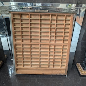 Puede incluir: Una bandeja de impresora de madera vintage con numerosos compartimentos pequeños. La bandeja es de madera de color claro y tiene forma rectangular. Se coloca sobre un fondo espejado con estatuas decorativas a ambos lados.