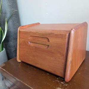 May include: A rectangular wooden bread box with a warm brown finish and a smooth, natural wood grain. The front panel features a horizontal recessed handle, and the side panels have rounded edges, creating a clean, solid, and functional storage container.