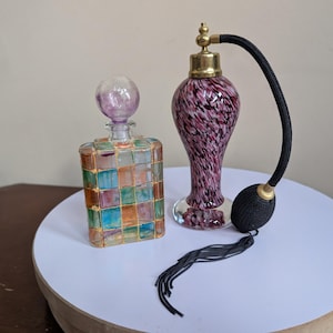 May include: A rectangular glass perfume bottle with a grid of multicolored squares and a round purple stopper, alongside a teardrop-shaped purple and white speckled glass perfume atomizer with a gold-tone metal top and a black fabric bulb pump.