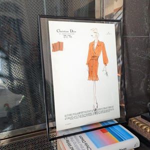 Könnte beinhalten: Gerahmte Modeillustration einer Frau in einem orangefarbenen Kleid mit Gürtel, von Christian Dior. Die Zeichnung ist in schwarzer Tinte mit orangefarbenen Aquarellakzenten gehalten. Der Rahmen ist schwarz und ruht auf einem Stapel Bücher.