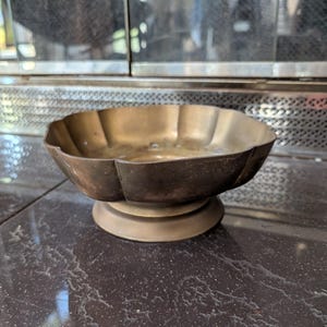 May include: A vintage brass bowl with a scalloped rim and a pedestal base. The bowl has a dark, aged patina, suggesting it is an antique. The bowl is likely used for decorative purposes or as a small serving dish.