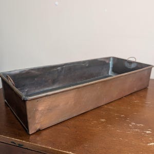 May include: A rectangular copper baking pan with a weathered, metallic finish. The pan features straight, vertical sides and a flat base, with small, thin metal loops attached to the short ends to serve as handles for lifting and carrying.