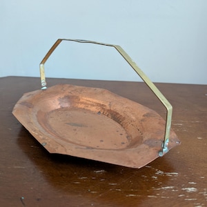 May include: An oval-shaped copper tray with a hammered texture and a flat, angled rim. The tray features a tall, thin, brass-colored metal handle attached to the sides with small rivets, allowing the handle to pivot upward from the base.