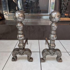 Puede incluir: Un par de morillos de chimenea de metal plateado con un poste vertical rematado por un remate esférico, una columna central con dos elementos decorativos redondeados más pequeños y una base con volutas y detalles tallados.