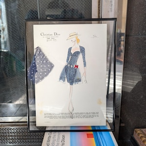 Könnte beinhalten: Gerahmte Christian Dior Modeillustration. Das Kunstwerk zeigt eine Skizze einer Frau in einem blauen Kleid mit roter Schleife, passendem Hut und einem abgetrennten Stoffmuster. Der Rahmen ist schwarz und das Kunstwerk wird auf weißem Hintergrund gezeigt.