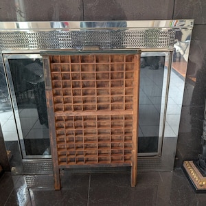 Puede incluir: Una bandeja de impresora de madera vintage con numerosos compartimentos pequeños. La bandeja está hecha de madera envejecida con un acabado oscuro y está soportada por dos patas. El fondo incluye una chimenea con marco de acero inoxidable y estatuas decorativas.