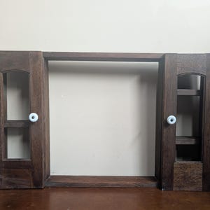 May include: A dark brown wooden wall-mounted cabinet frame featuring two side sections with open rectangular cutouts and small white ceramic knobs. The structure includes a central open space and horizontal top and bottom connecting beams.