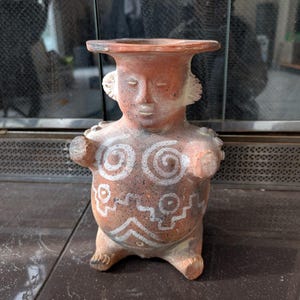 May include: A terracotta-colored ceramic figurine with a human-like form. The figure has a wide, flat head and is decorated with white spiral and geometric patterns on the chest and abdomen. The figurine has small arms and legs.