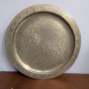 May include: A large, circular brass tray featuring an intricate, hand-engraved geometric star pattern in the center and a repeating floral motif along the wide, raised rim. The metal surface has a muted, aged gold-toned finish.
