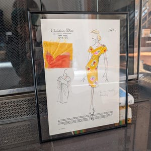 Könnte beinhalten: Gerahmte Christian Dior Modezeichnung aus Paris 1956. Die Skizze zeigt ein gelbes Kleid mit floralen Akzenten, einen Stoffmuster und weitere Design-Details. Das Kunstwerk ist in einem schwarzen Rahmen ausgestellt.