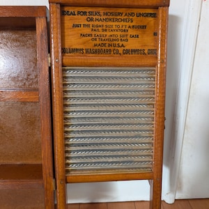 Op de afbeelding: Een vintage houten wasbord met een glazen schrobvlak. Het wasbord heeft een houten frame met tekst die luidt "IDEAL FOR SILKS, HOSIERY AND LINGERIE" en "COLUMBUS WASHBOARD CO., COLUMBUS, OHIO".