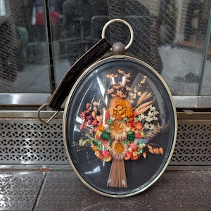 May include: Oval framed floral arrangement with a glass cover. The bouquet features dried flowers in shades of orange, red, yellow, and green. A dark brown leather strap is attached to the top. The frame is metallic.