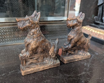 May include: Pair of vintage brown dog figurines, resembling Scottish Terriers, sitting on small wooden fences. The dogs have detailed fur texture and collars. Decorative home decor.