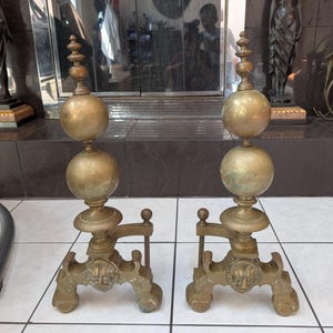Puede incluir: Un par de morillos de chimenea de latón a juego con un diseño vertical que presenta dos remates esféricos apilados sobre una base. La base de cada morillo está decorada con un motivo de cabeza de león esculpida y apoyada sobre cuatro patas de garra.