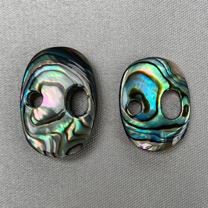 Può includere: Due bottoni in madreperla di abalone iridescenti con due fori ciascuno. I bottoni sono di forma ovale e hanno una gamma di colori verde, blu e viola.