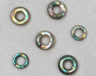 May include: Nine iridescent abalone shell rings in various sizes. The rings are arranged in a semi-circle on a light gray fabric background.