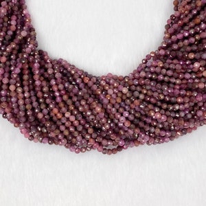 Könnte beinhalten: Nahaufnahme einer Halskette aus kleinen, runden, facettierten Perlen in Burgunder- und Mauve-Tönen. Die Perlen sind aufgefädelt und erzeugen einen geschichteten Effekt. Die Halskette wird vor einem weißen Hintergrund präsentiert.