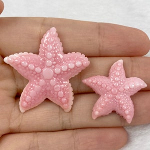 Op de afbeelding: Twee roze zeesterdecoraties met een textuur. De grotere zeester heeft een diameter van ongeveer 5 cm, terwijl de kleinere ongeveer 2,5 cm meet. Beide hebben een lichtroze kleur met donkerroze details.