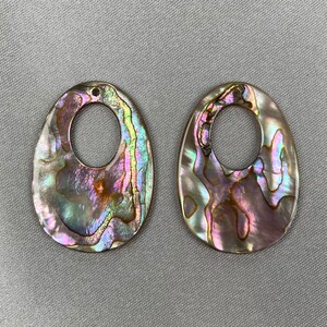 Könnte beinhalten: Zwei schillernde Abalone-Muschel-Anhänger mit ovaler Form und einem zentralen Loch. Die Muscheln zeigen eine Farbpalette aus Rosa, Grün und Blau mit einem perlmuttartigen Glanz. Ideal für die Schmuckherstellung.